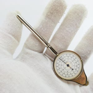 May include: A vintage silver-toned circular slide rule with a rotating dial and a long handle. The dial features black numbers and markings on a white background. The slide rule is held against a white background.