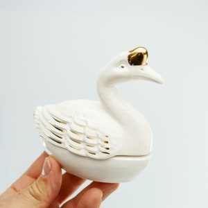 Può includere: Scatola portaoggetti in ceramica bianca a forma di cigno con dettagli dorati. Il cigno ha un design dettagliato e un coperchio con cerniera.