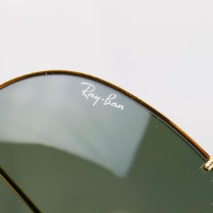 May include: Close-up of Ray-Ban sunglasses. The lenses are a gradient green, and the frame is gold. The Ray-Ban logo is visible in white on the upper left lens. The sunglasses are set against a white background.