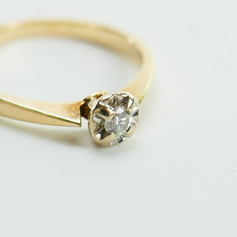 K&ouml;nnte beinhalten: Ein einfacher Goldring mit einem einzigen, kleinen, runden Diamanten in einer Krappenfassung.