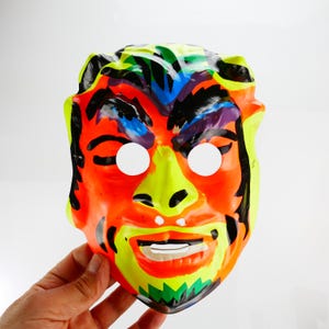 May include: A colorful Halloween mask with a smiling face. The mask features bright orange, yellow, green, blue, and black paint. The eye holes are white. The mask is held by a hand.