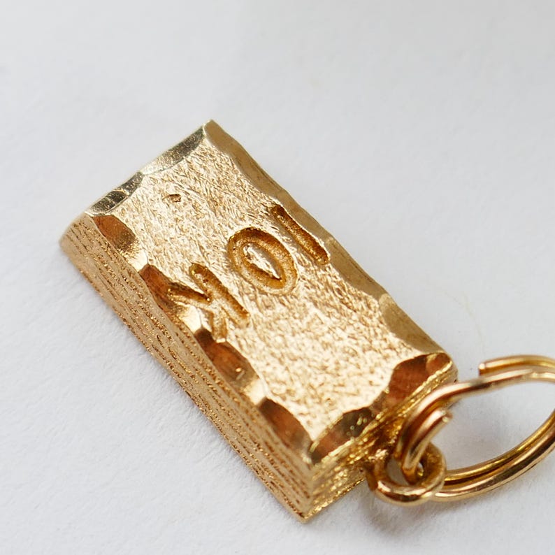 May include: A gold-colored metal charm in the shape of a bar with the word "MOM" engraved on it. The charm is attached to a gold-colored metal key ring.
