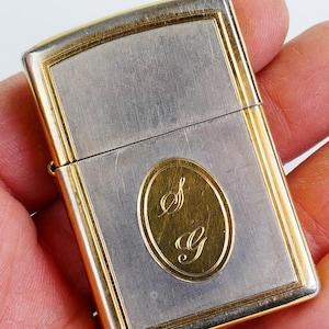 May include: A silver and gold-toned lighter with an oval gold emblem featuring the letters "S" and "G". The lighter has a rectangular shape with gold trim and a brushed silver centre.