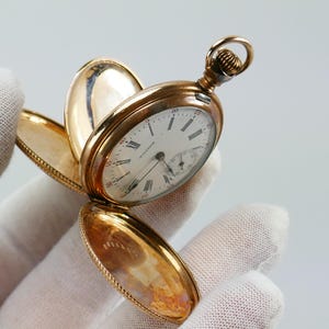 May include: A gold pocket watch with an open face, revealing a white clock face with black Roman numerals and delicate hands. The watch case is a warm gold color, and the watch is held open, showcasing its intricate design and the word "WALTHAM".