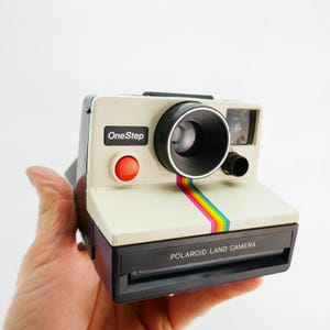 Peut inclure: Appareil photo Polaroid OneStep Land de couleur crème avec un objectif noir et un bouton rouge. Une bande arc-en-ciel orne l'avant. Les textes "OneStep" et "POLAROID LAND CAMERA" sont visibles.