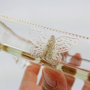 May include: Clear glass bottle with a decorative butterfly design. The butterfly is detailed with textured wings and body. The bottle has a gold trim along the edge.
