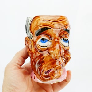 Può includere: Una tazza in ceramica vintage a forma di testa di uomo, con capelli marroni, neri e bianchi, occhi blu e un colletto rosa. La tazza è tenuta in mano su sfondo bianco. La tazza ha un volto dettagliato ed espressivo.