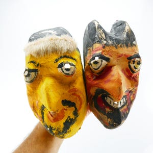 May include: Two handmade, folk art masks. One mask is yellow with a fur trim and black accents. The other mask is orange and black with a sinister expression. Both masks have large, expressive eyes and are held against a white background.
