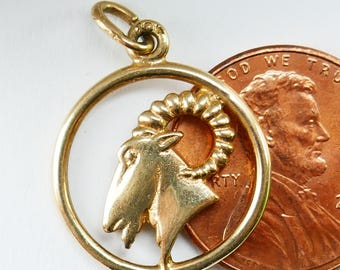 May include: Gold-toned pendant charm featuring a detailed depiction of a ram's head with curled horns, set within a circular frame. The charm is approximately the size of a US penny.