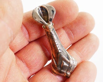 May include: A silver pendant with a distinctive, elongated shape. The pendant has a flared top and a tapering body with geometric patterns. A small gold-coloured accent is visible near the top. The pendant is held in a hand against a white background.