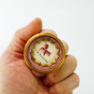 Op de afbeelding: Een klein, rond, goudkleurig reliekhouder met een roze lint en de tekst "S.M. Bernardae" erin.
