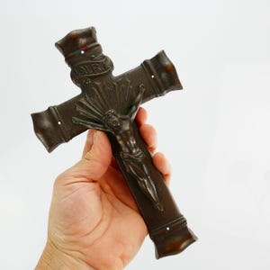 Könnte beinhalten: Ein bronzefarbenes Kruzifix mit einer detaillierten Darstellung von Jesus Christus. Das Kreuz hat dekorative Kanten und die Inschrift "I.N.R.I." oben. Die Figur Christi ist zentral positioniert, mit Strahlen, die von hinter seinem Kopf ausgehen.