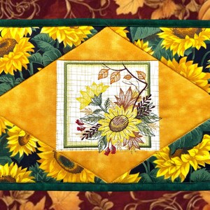 May include: Rectangular quilted placemat featuring a sunflower design. The placemat has a dark green border and a central diamond shape in yellow. The diamond shape surrounds a square with a floral bouquet and a grid pattern.