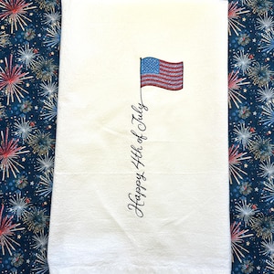 May include: White kitchen towel with an embroidered American flag and the words "Happy 4th of July" in black script. The towel is displayed on a blue background with a repeating pattern of red, white, and blue fireworks.