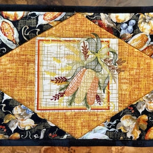 May include: A rectangular, quilted placemat with a black border. The center features a diamond-shaped design in orange fabric, framing a square with embroidered corn and wheat. The background is black with fall-themed motifs.