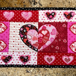May include: A pink and red quilted heart-shaped coaster with a black and white polka dot border. The coaster is made of fabric and has a variety of heart designs embroidered on it.