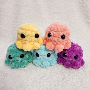 May include: Five crocheted octopus plushies in different colors: teal, light blue, pink, yellow, and purple. Each octopus has black eyes and a textured, fuzzy surface.