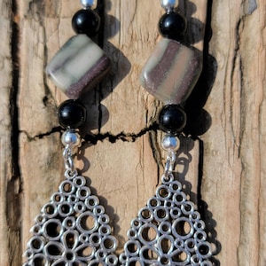 May include: A pair of dangle earrings with a silver-tone filigree teardrop design. The earrings feature black and silver beads, and square-shaped stones with brown, tan, and green stripes. The earrings hang from silver-tone hooks.