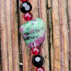 Puede incluir: Primer plano de un collar con una gran cuenta de piedra verde y morada, rodeada de cuentas rojas y negras más pequeñas. El collar está ensartado en un alambre fino, sobre un fondo de madera.