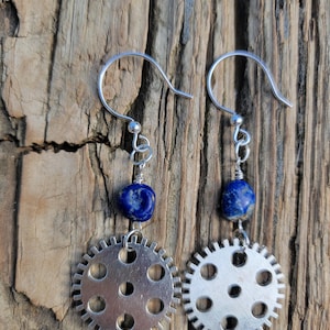 Pode incluir: Brincos prateados com design steampunk. Cada brinco apresenta um pingente em forma de engrenagem com furos, combinado com uma conta azul de lápis-lazúli. Os brincos pendem de ganchos prateados.