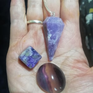 May include: A collection of purple and white gemstones, including a faceted pendulum on a silver chain, a diamond-shaped stone, and an oval stone. The stones are held in a hand.