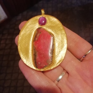 May include: A gold oval pendant with a pink and white stone set in the center. A small purple stone is set at the top of the pendant.