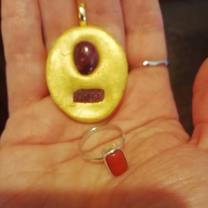 May include: A gold pendant with a red gemstone set in a circular frame. A silver ring with a red rectangular gemstone.