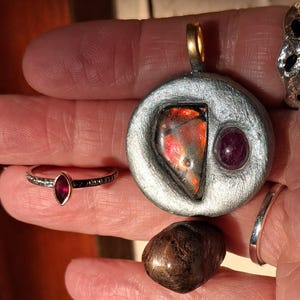 Może przedstawiać: Srebrny wisiorek z dużą, nieregularną, wielokolorową kamieniem i mniejszym, okrągłym, czerwonym kamieniem. Widoczny jest również srebrny pierścionek z czerwonym kamieniem w kształcie markizy i brązowy, polerowany kamień.