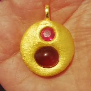 Może przedstawiać: Złoty wisiorek z dwoma czerwonymi kamieniami szlachetnymi. Większy kamień jest okrągły, a mniejszy kwadratowy.