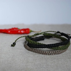 Può includere: Tre braccialetti intrecciati in nero, verde e marrone. I braccialetti sono realizzati in cordoncino intrecciato e hanno un design semplice e minimalista.