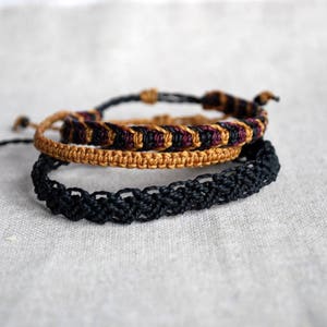 May include: Three handmade macrame bracelets in varying colors. The top bracelet is black with a tan knot, the middle bracelet is tan with black and burgundy accents, and the bottom bracelet is black.