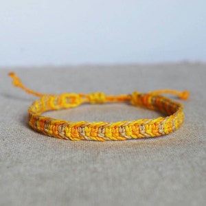 Könnte beinhalten: Ein handgefertigtes Armband mit einem gewebten Design in Gelb- und Beigetönen. Das verstellbare Armband besteht aus geflochtenem Kordel und hat ein schlichtes, aber stilvolles Aussehen. Das Armband ist für den täglichen Gebrauch geeignet.