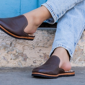May include: Brown leather slip-on shoes with a textured finish. The shoes have a black sole and a light brown trim. The design features a rounded toe and a low-profile heel. The shoes are shown on a person wearing blue jeans.