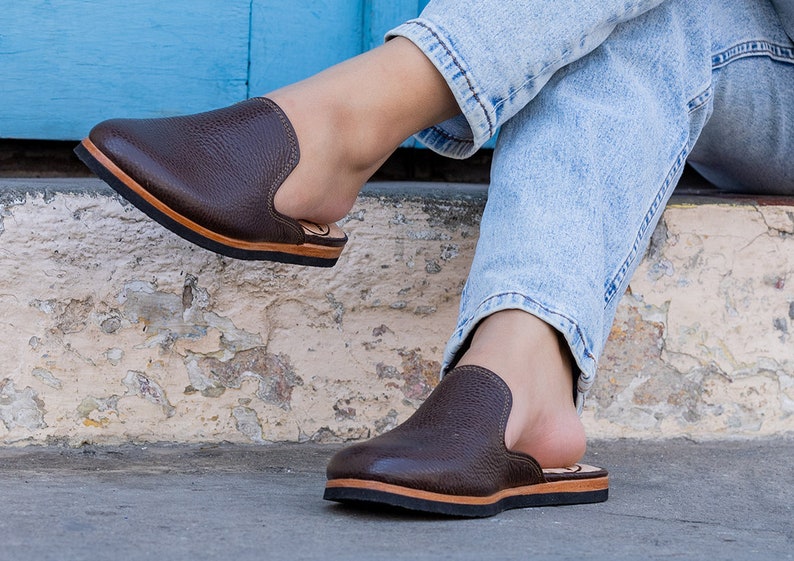 May include: Brown leather slip-on shoes with a textured finish. The shoes have a black sole and a light brown trim. The design features a rounded toe and a low-profile heel. The shoes are shown on a person wearing blue jeans.