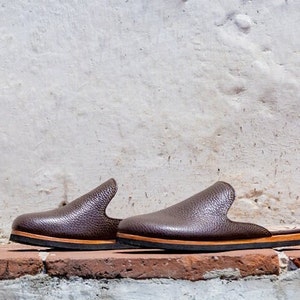 May include: A pair of brown leather slip-on shoes with a textured surface. The shoes have a rounded toe and a low heel. The soles are black with a thin tan stripe. The shoes are displayed on a red brick surface against a white wall.