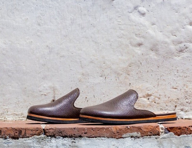 May include: A pair of brown leather slip-on shoes with a textured surface. The shoes have a rounded toe and a low heel. The soles are black with a thin tan stripe. The shoes are displayed on a red brick surface against a white wall.
