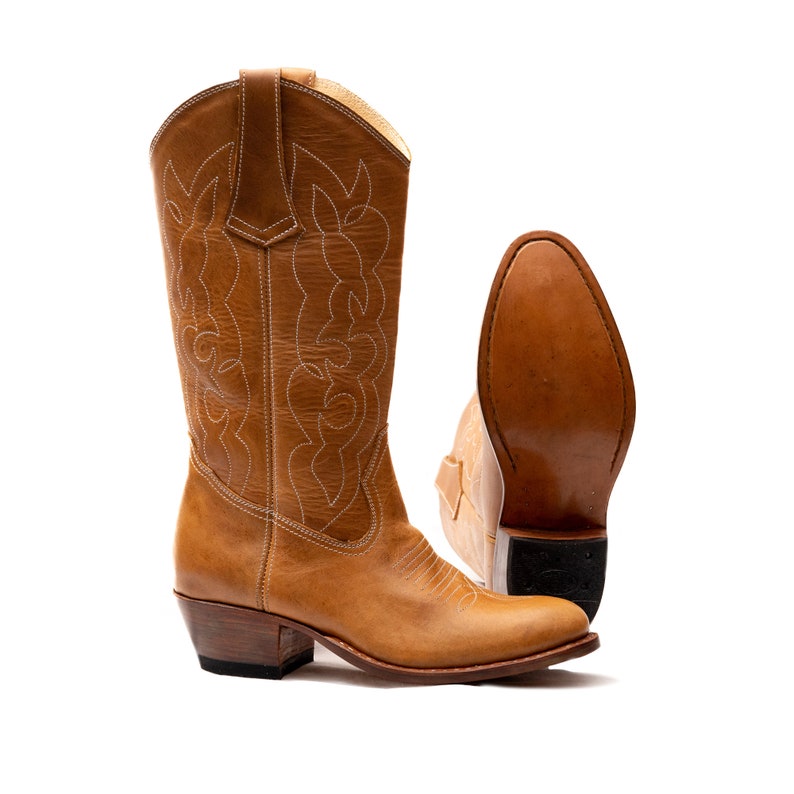 May include: A pair of brown leather cowboy boots with white stitching. The boots have a pointed toe and a low heel. One boot is standing upright, while the other is lying on its side, showing the sole.
