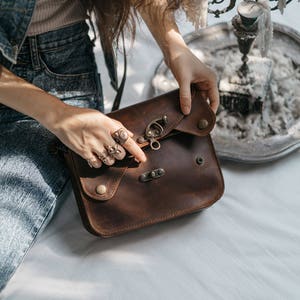 May include: A brown leather satchel with a flap closure and metal hardware. The bag features a buckle clasp and decorative rivets. The satchel is held by a person wearing multiple rings.
