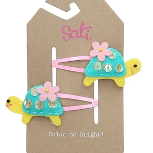 以下が含まれることがあります： ピンクのヘアクリップ2つに、ターコイズブルーのフェルトでできた黄色い頭とピンクの花のついたカメが飾られています。カメの甲羅にはスパンコールが施されています。パッケージには「Sati」と「Color me bright!」の文字が書かれています。