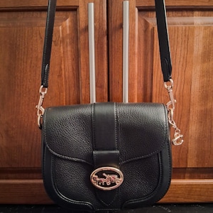 May include: A black leather crossbody bag with a pebbled texture, featuring a gold-tone metal clasp on the front flap that depicts a horse and carriage logo. The bag includes a matching black leather shoulder strap attached with gold-tone hardware.
