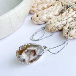 May include: A silver necklace featuring a teardrop-shaped pendant made from a polished agate stone. The agate displays bands of brown, white, and gray, with a crystal-filled center. The pendant is set in a silver bezel and hangs from a delicate silver chain.
