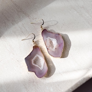 May include: A pair of agate earrings with a gold-colored trim. The earrings have a light purple hue with a white crystal center. The earrings are displayed on a white surface, with the sunlight casting a shadow.
