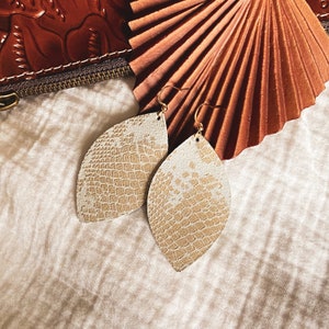May include: A pair of gold tone earrings with a light brown, textured, leaf-shaped design.