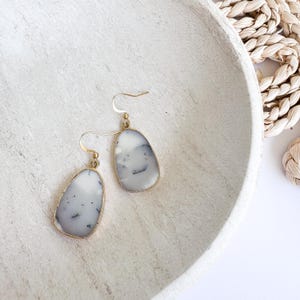 May include: A pair of dangle earrings with a gold-colored frame. The earrings feature a light gray stone with black and white marbling. The earrings are displayed on a white, textured surface.