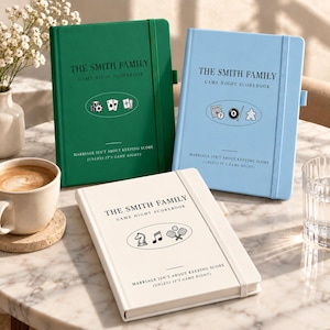 Puede incluir: Tres cuadernos de puntuación de tapa dura en verde, azul claro y crema, cada uno con cierre elástico, el texto THE SMITH FAMILY GAME NIGHT SCOREBOOK, iconos de juegos y la frase MARRIAGE ISN'T ABOUT KEEPING SCORE (UNLESS IT'S GAME NIGHT).