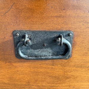 May include: Close-up of a dark, rectangular metal drawer pull with a curved handle, mounted on a wooden surface. The pull has a distressed, aged appearance, with visible wear and tear. The wood has a warm, reddish-brown tone.