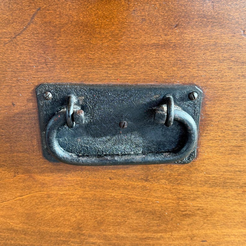 May include: Close-up of a dark, rectangular metal drawer pull with a curved handle, mounted on a wooden surface. The pull has a distressed, aged appearance, with visible wear and tear. The wood has a warm, reddish-brown tone.