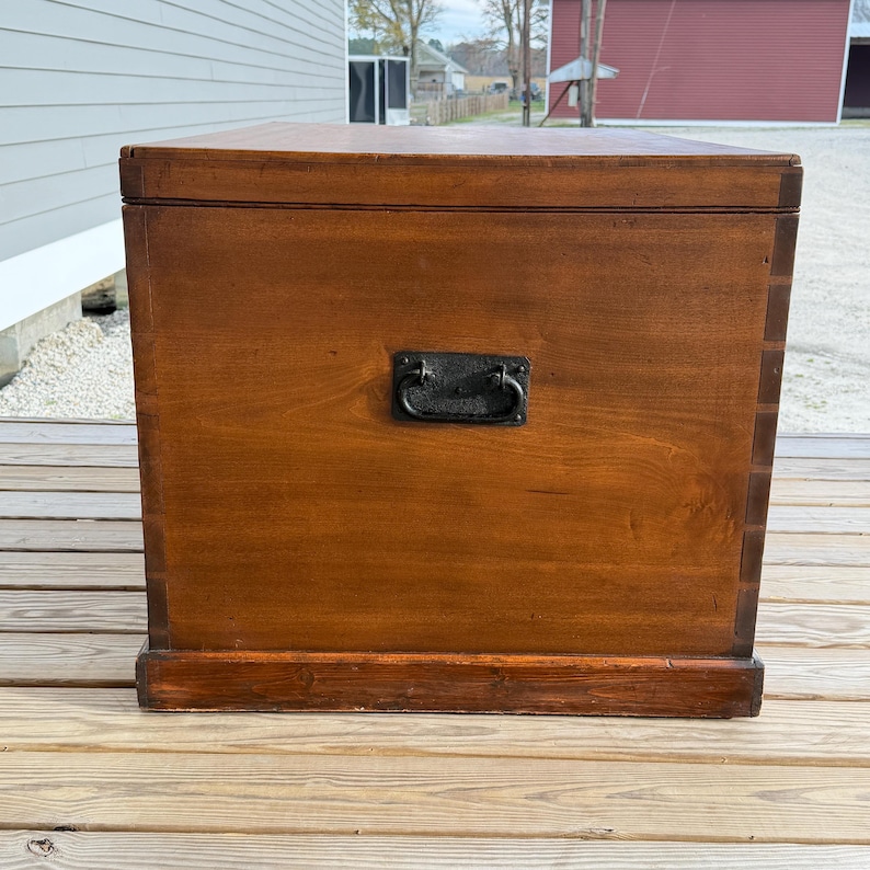 May include: A rectangular wooden chest with a dark brown finish. It features a black metal handle and is set on a wooden surface. The chest has a simple, classic design, suitable for storage.
