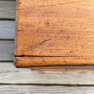 May include: Close-up of a wooden surface with a warm, reddish-brown hue. The wood grain is visible, with natural variations in color and texture. The corner of the wooden object is visible, showing the edge and the way the wood pieces are joined.