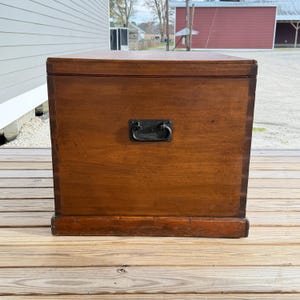 May include: A rectangular wooden chest with a dark brown finish. It features a metal handle and a base. The chest is likely used for storage and has a classic, vintage appearance. The wood grain is visible.